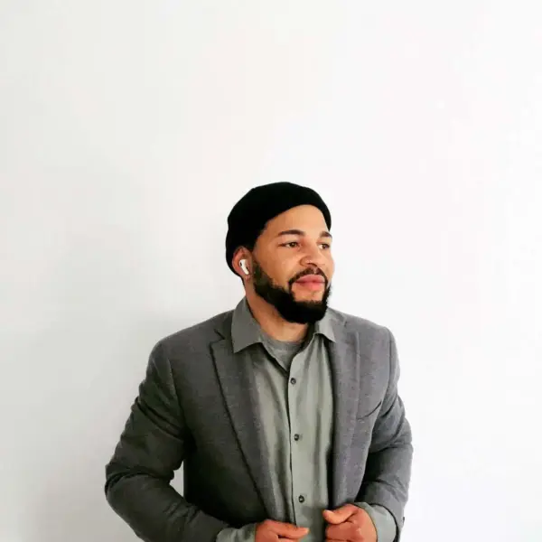 Mann im Business-Outfit mit schwarzen Beanie und AirPods vor weißer Wand.